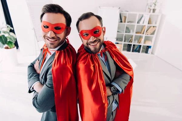 Blick aus der Vogelperspektive auf lächelnde gutaussehende Superunternehmer in Masken und Umhängen mit verschränkten Armen im Büro — Stockfoto