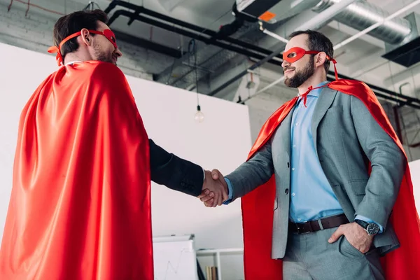 Low angle view of handsome super businessmen in masks and capes shaking hands in office — Stock Photo