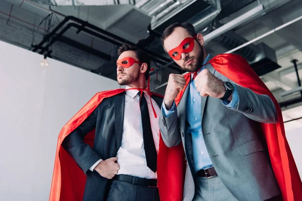 Vue en angle bas de beaux super hommes d'affaires en masques et capes montrant les poings au bureau — Photo de stock