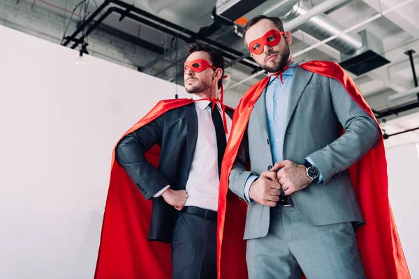 Visão de baixo ângulo de bonitos super empresários em máscaras vermelhas e capas no escritório — Fotografia de Stock