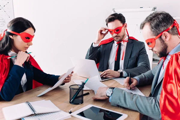 Nachdenkliche Superunternehmer in Masken und Umhängen, die im Büro arbeiten — Stockfoto