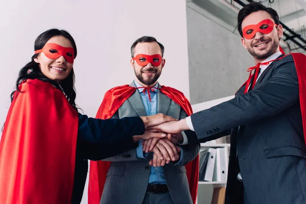 Super businesspeople in masks and capes putting hands together in office — Stock Photo