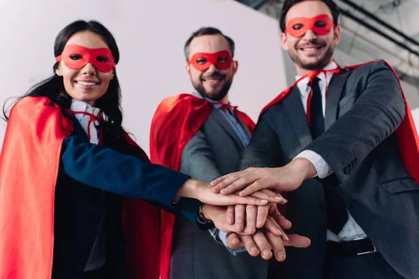 Vista basso angolo di super uomini d'affari in maschere e mantelli mettendo le mani insieme in ufficio — Foto stock