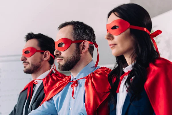 Vista lateral de super empresarios en máscaras y capas mirando hacia otro lado en la oficina - foto de stock