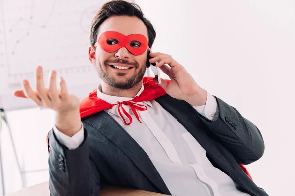 Guapo super hombre de negocios en máscara y capa hablando por teléfono inteligente en la oficina - foto de stock