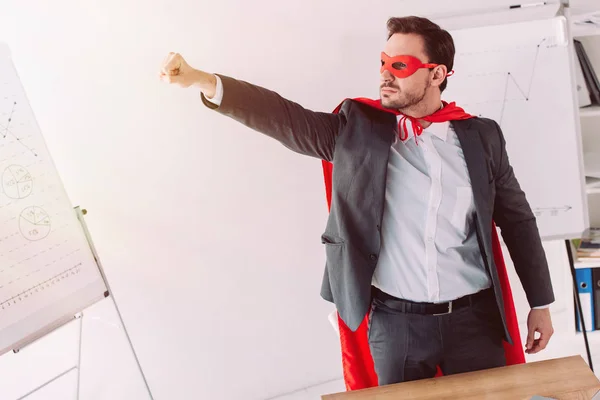 Handsome super businessman in mask and cape standing with hand up in office — Stock Photo