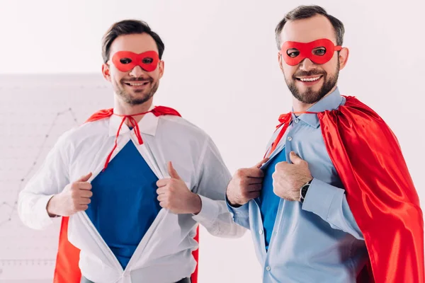 Beaux super hommes d'affaires en masques et capes montrant chemises bleues au bureau — Photo de stock