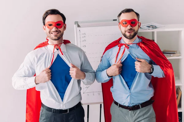 Souriant beaux super hommes d'affaires en masques et capes montrant chemises bleues au bureau — Photo de stock