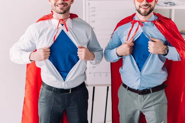 Imagem cortada de super empresários em máscaras e capas mostrando camisas azuis no escritório — Fotografia de Stock