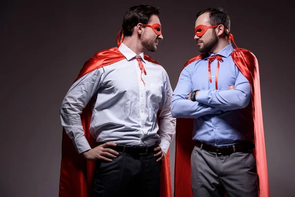 Guapos súper hombres de negocios con máscaras y capas mirándose aislados en gris - foto de stock