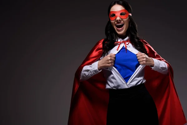 Laughing attractive super businesswoman in red cape and mask showing blue shirt isolated on black — Stock Photo