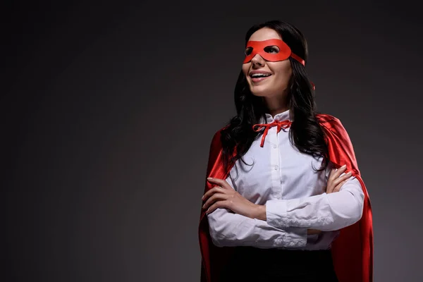 Souriant attrayant super femme d'affaires en cape rouge et masque debout avec les bras croisés isolés sur noir — Photo de stock