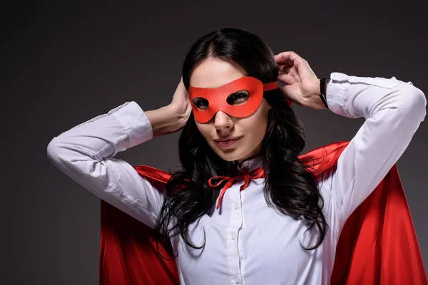 Attractive super businesswoman in red cape tying mask isolated on black — Stock Photo