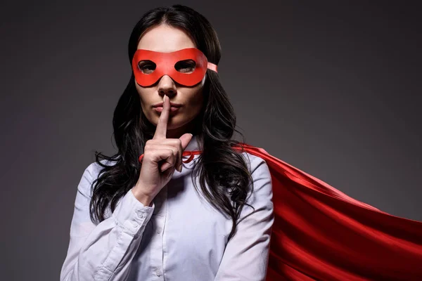 Attractive super businesswoman in red cape and mask showing silence gesture isolated on black — Stock Photo