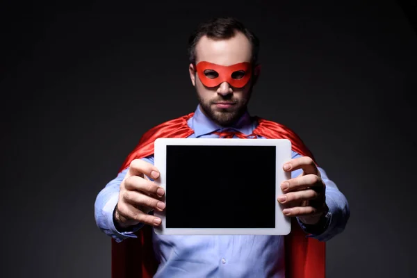 Красивый супер бизнесмен в маске и плаще показывает планшет изолирован на черном — стоковое фото