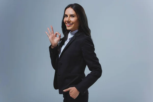 Smiling attractive businesswoman showing okay gesture isolated on grey — Stock Photo