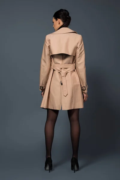 Vista trasera de mujer en pantimedias negras y zanja beige sobre fondo oscuro - foto de stock