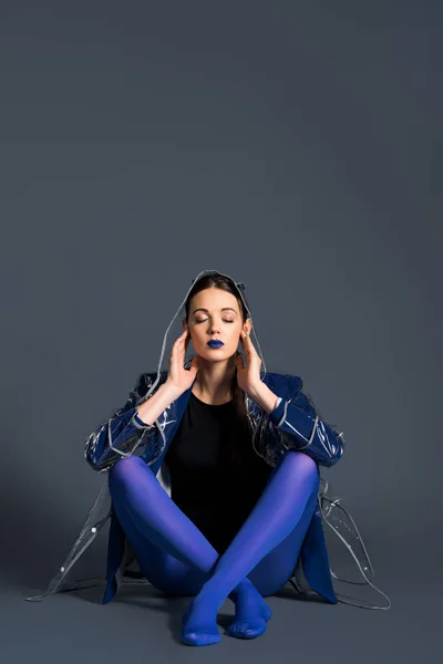 Девушка в прозрачном плаще и синих колготках сидит на тёмном фоне — стоковое фото