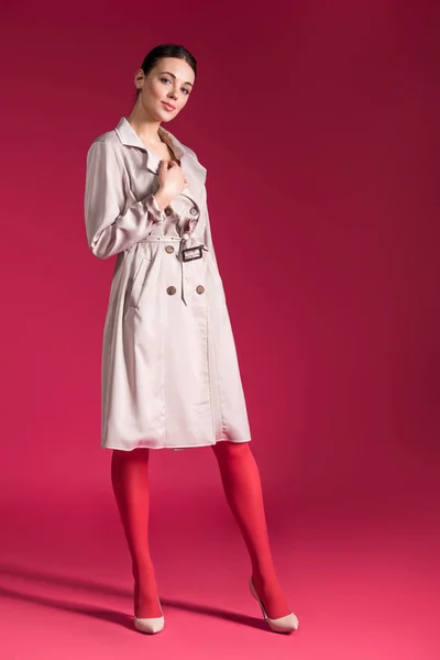 Улыбающаяся девушка в бежевом окопе и на высоких каблуках на красном фоне — стоковое фото