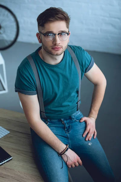 Hochwinkelaufnahme eines gutaussehenden jungen Mannes mit Brille, der auf einem Tisch sitzt und in die Kamera blickt — Stockfoto
