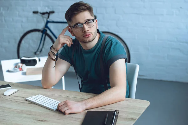 Handsome pensive freelancer in eyeglasses looking away while sitting at workplace — Stock Photo