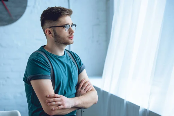 Joven pensativo de pie con los brazos cruzados y mirando a la ventana - foto de stock