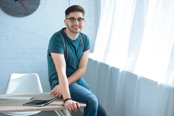 Schöner junger Mann mit Brille lächelt in die Kamera, während er im Home Office auf dem Tisch sitzt — Stockfoto