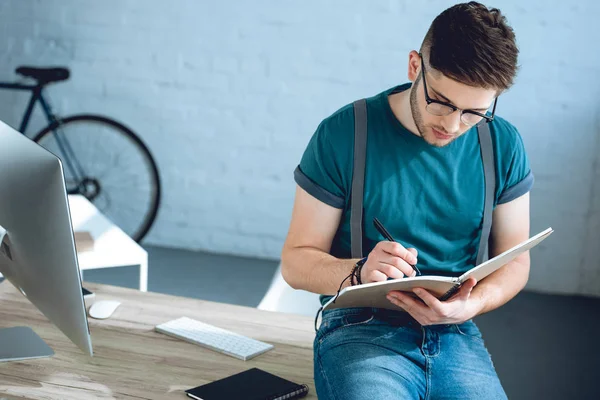 Guapo joven freelancer en gafas tomando notas en el lugar de trabajo - foto de stock