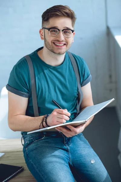 Bello giovane uomo in occhiali prendere appunti e sorridere alla macchina fotografica — Foto stock