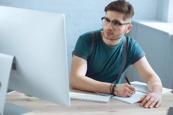 Schöner junger Freiberufler mit Brille, der Notizen im Notizbuch macht, während er mit dem Desktop-Computer arbeitet — Stockfoto