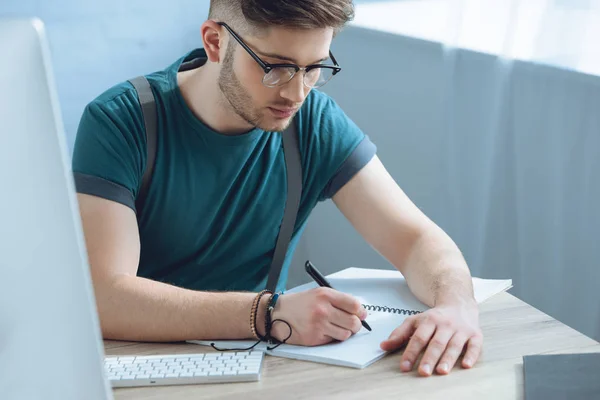 Jeune homme concentré dans les lunettes prenant des notes tout en travaillant au bureau à domicile — Photo de stock