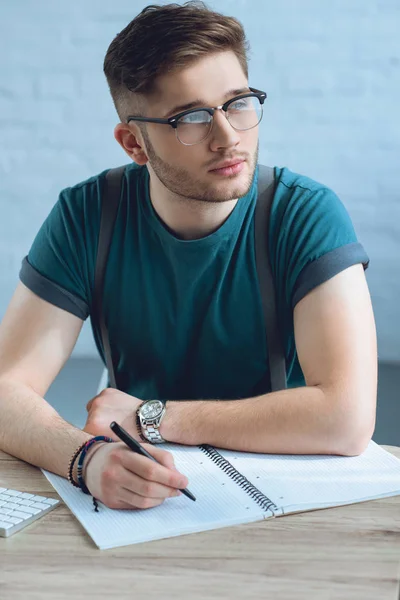 Pensive young man in eyeglasses looking away while writing in notebook — Stock Photo