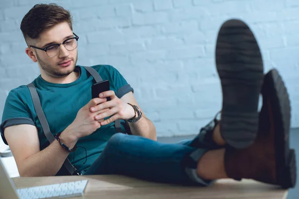 Handsome yougn man in eyeglasses sitting with feet on table and using smartphone — Stock Photo