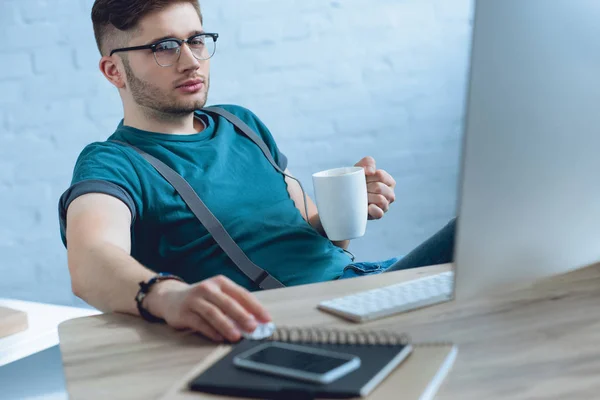 Joven y en gafas con taza y el uso de la computadora de escritorio - foto de stock