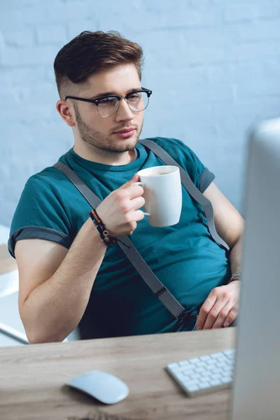 Joven con anteojos bebiendo café y trabajando con computadora de escritorio en casa oficina - foto de stock