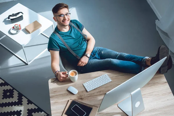 Vue grand angle de beau jeune homme en lunettes souriant à la caméra tout en étant assis sur le lieu de travail au bureau à la maison — Photo de stock