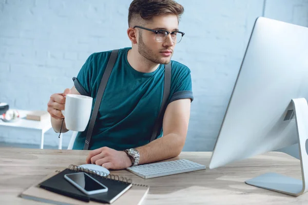 Guapo joven freelancer en gafas con taza y trabajando con computadora de escritorio - foto de stock