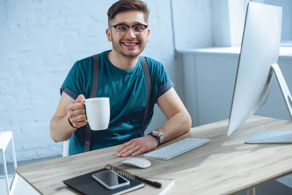 Schöner junger Mann mit Tasse und lächelnd in die Kamera, während er mit dem Desktop-Computer arbeitet — Stockfoto