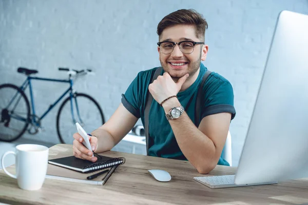 Allegro giovane uomo con gli occhiali sorridente alla fotocamera mentre lavorava a casa ufficio — Foto stock