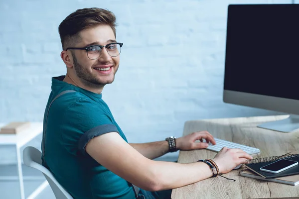 Schöner junger Mann mit Brille, der mit einem Desktop-Computer arbeitet und in die Kamera lächelt — Stockfoto