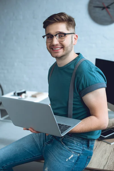 Bel giovane uomo con gli occhiali che tiene il computer portatile e sorride alla fotocamera — Foto stock