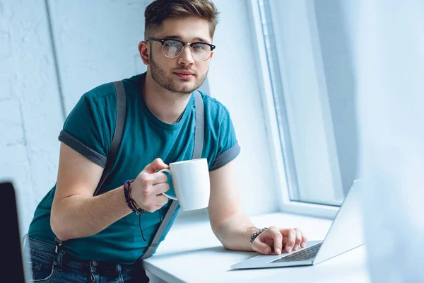 Junger Mann mit Brille hält eine Tasse Kaffee in der Hand und schaut in die Kamera, während er zu Hause den Laptop benutzt — Stockfoto