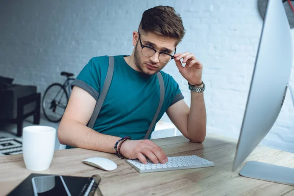 Guapo joven freelancer ajustando gafas y mirando a la cámara mientras trabaja con computadora de escritorio - foto de stock