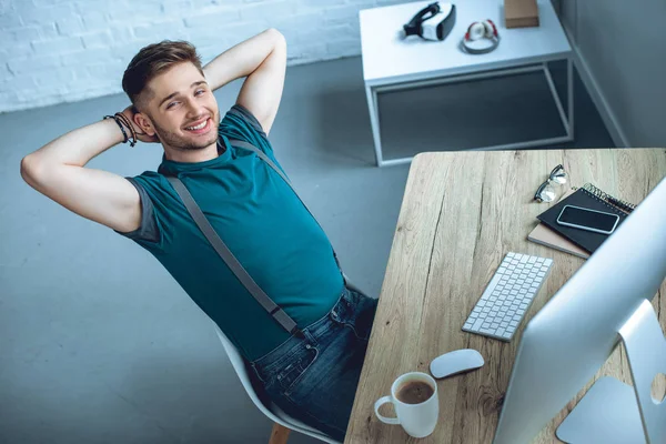 High angle view of handsome young freelancer sitting with hands behind head and smiling at camera — Stock Photo