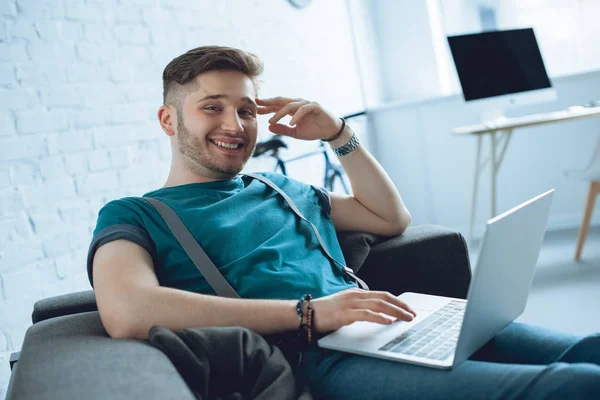Schöner junger Mann lächelt in die Kamera, während er zu Hause Laptop auf dem Sofa benutzt — Stockfoto