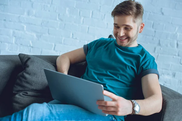 Sorrindo jovem usando laptop no sofá em casa — Fotografia de Stock