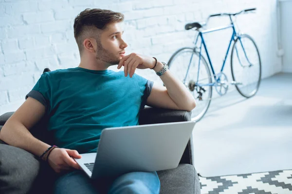 Schöner junger Mann benutzt Laptop und schaut weg, während er zu Hause sitzt — Stockfoto