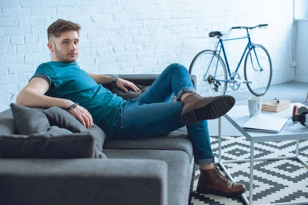 Schöner junger Mann sitzt auf der Couch und schaut zu Hause in die Kamera — Stockfoto
