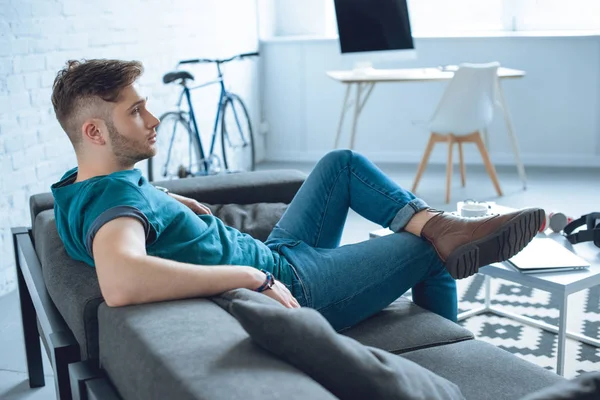 Vue latérale de beau jeune homme assis sur le canapé et regardant loin à la maison — Photo de stock