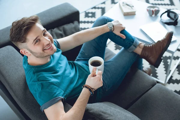 Vista de ángulo alto de joven guapo bebiendo café y sonriendo a la cámara en casa - foto de stock